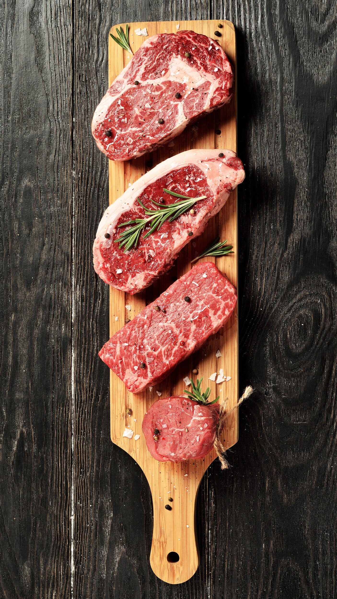 beef_mixed_ribeye_newyork_chuckflap_tenderloin Fresh cuts of marbled beef steaks arranged on a wooden cutting board, garnished with rosemary and black peppercorns, perfect for grilling or cooking.