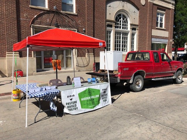 Outdoor farmers market setup featuring a red pickup truck and a vendor booth under a red canopy, displaying signs for pasture-raised poultry and Hidden Hollow products, with a checkered tablecloth and various items on the table.
