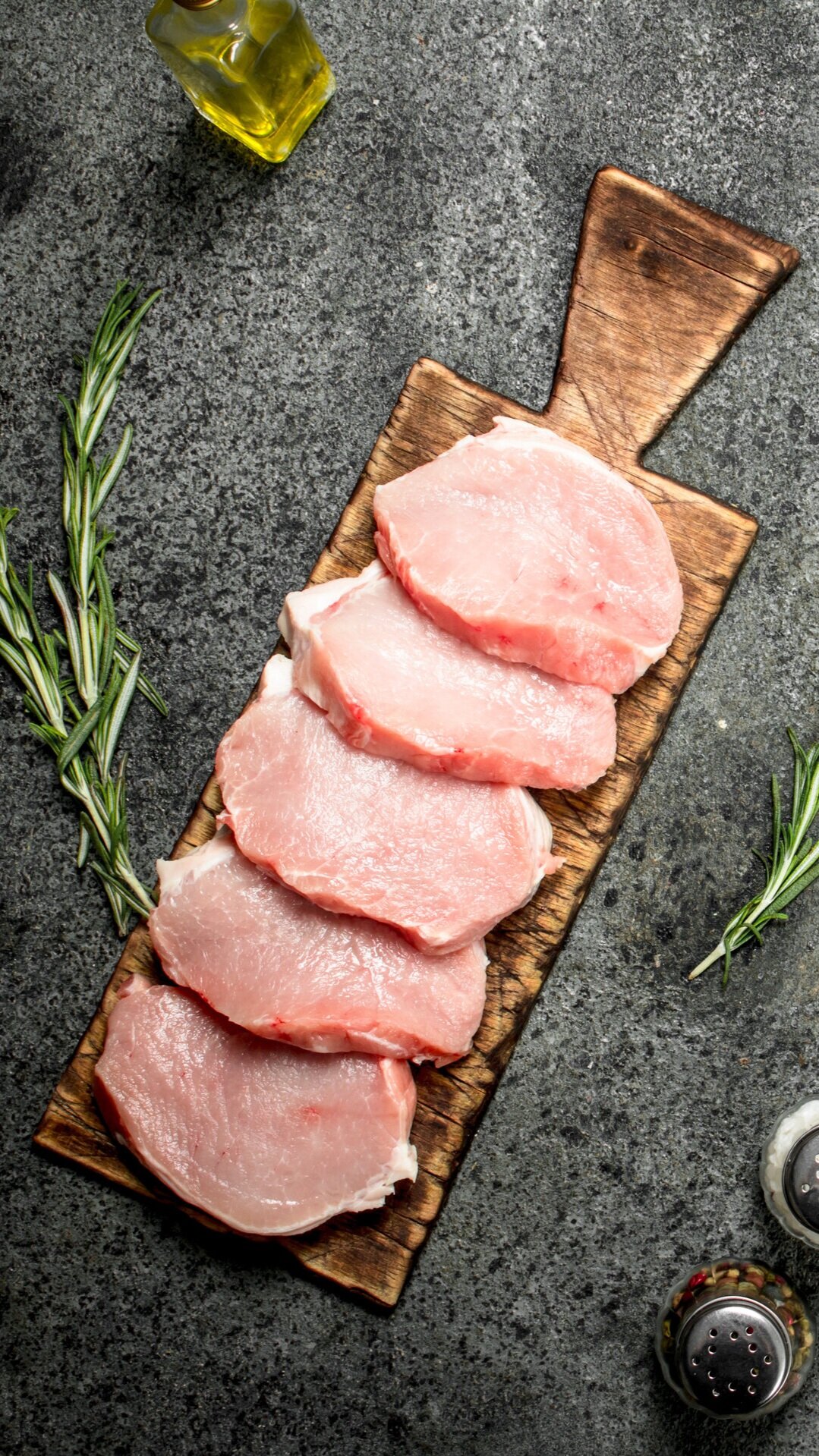 pork_chops_boneless_sliced_steaks Pork chops arranged on a wooden cutting board alongside fresh rosemary, olive oil, and salt and pepper shakers, set against a dark stone background.