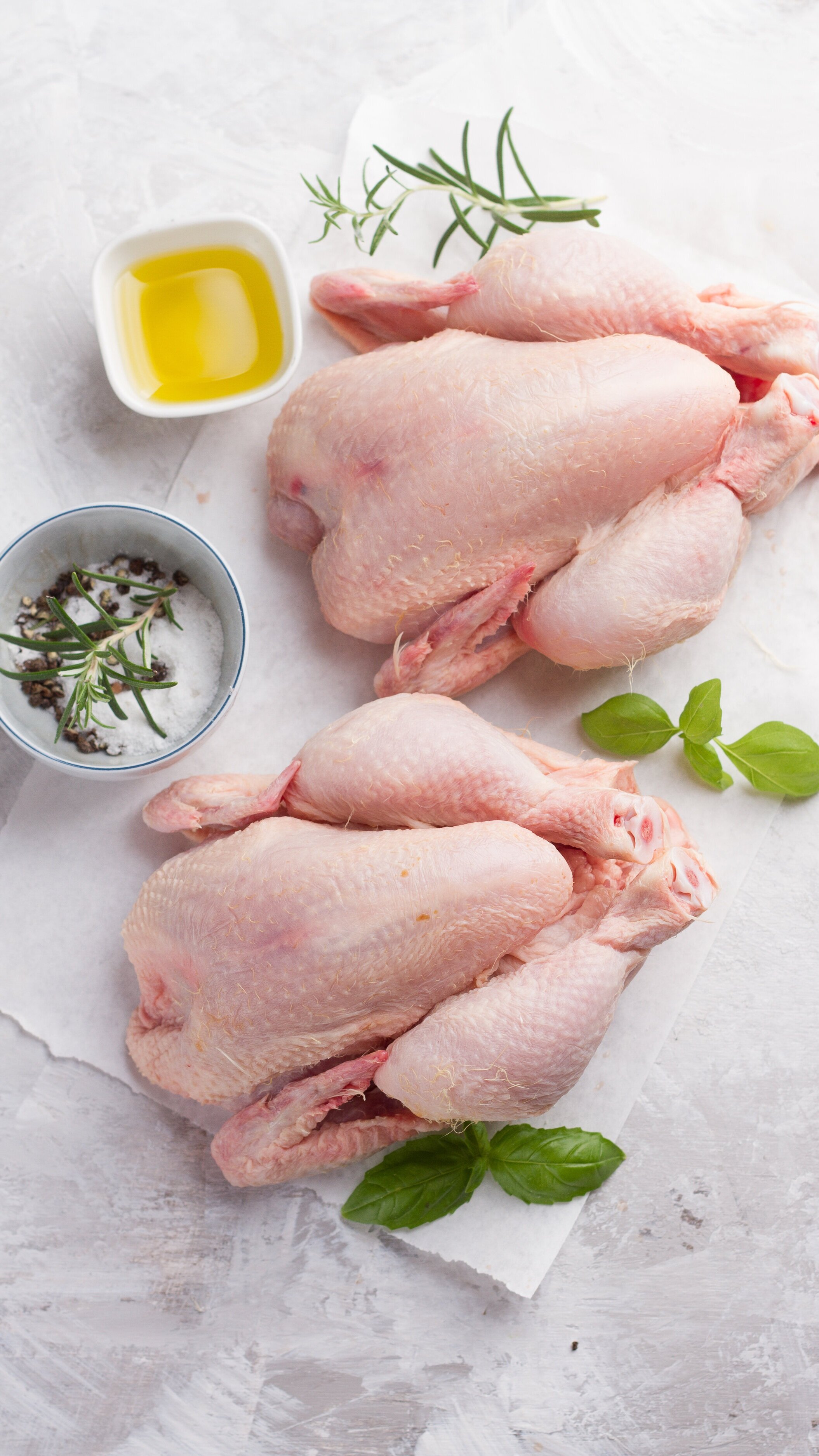 turkey_chickens_whole_marblewhitebackground Two raw whole chickens on a white surface, accompanied by a small bowl of salt and pepper, fresh rosemary, and a dish of olive oil, showcasing ingredients for a flavorful meal preparation.